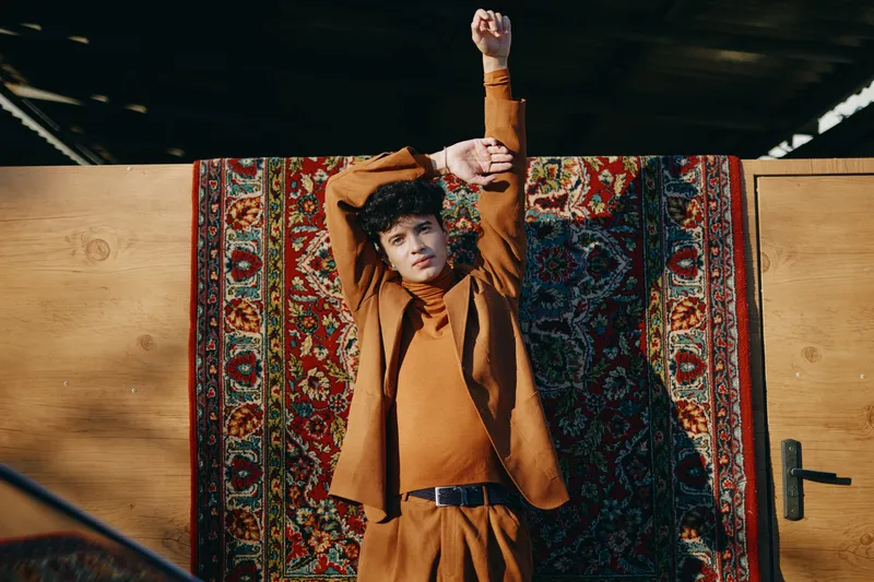 Модный мужчина-модель, одетый в оранжевый костюм, позирует с поднятыми руками на фоне красочного узорчатого ковра на деревянном полу