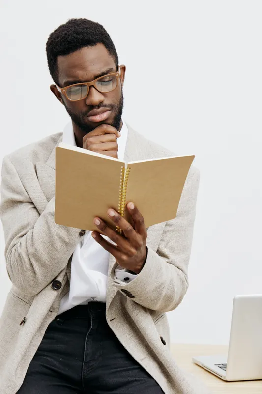Thoughtful young African American man with black hair, glasses, wearing a beige blazer and white shirt, reading a notebook while sitting at a desk against a plain white background in a modern office e