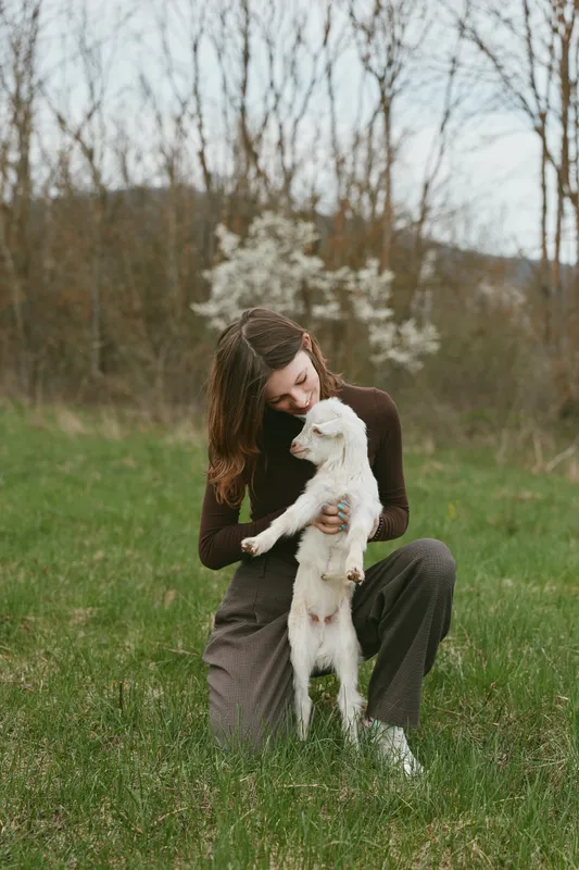 Young woman with long hair kneels on grass holding a white baby goat in her arms in a natural outdoor setting during early spring.