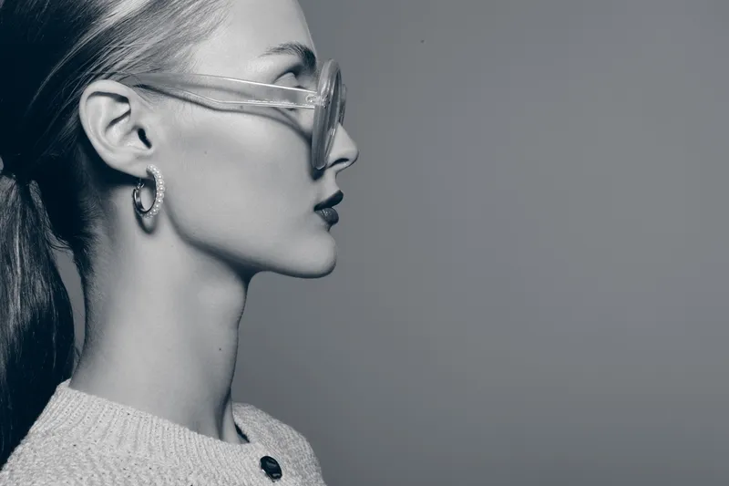 Profile of a young Caucasian woman with pale skin and glasses looks forward against grey background. Concept of fashion, elegance, and minimalism in monochrome style.
