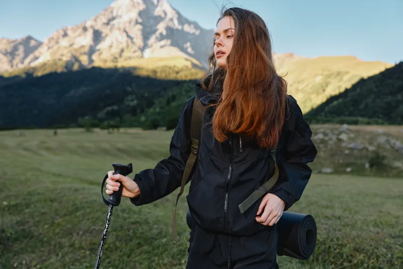 A rugged outdoor scene shows a young woman hiker moving along a hillside trail with a trekking pole. She wears a dark jacket and carries a mat, ready for exploration.