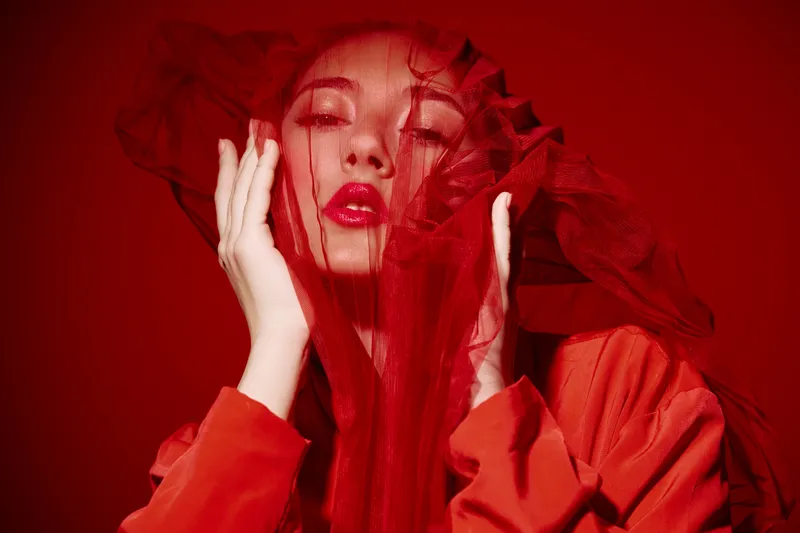 Creative beauty portrait of woman with bright red makeup and dramatic tulle veil against red background. Concept of fashion, color contrast, and artistic expression in studio light.