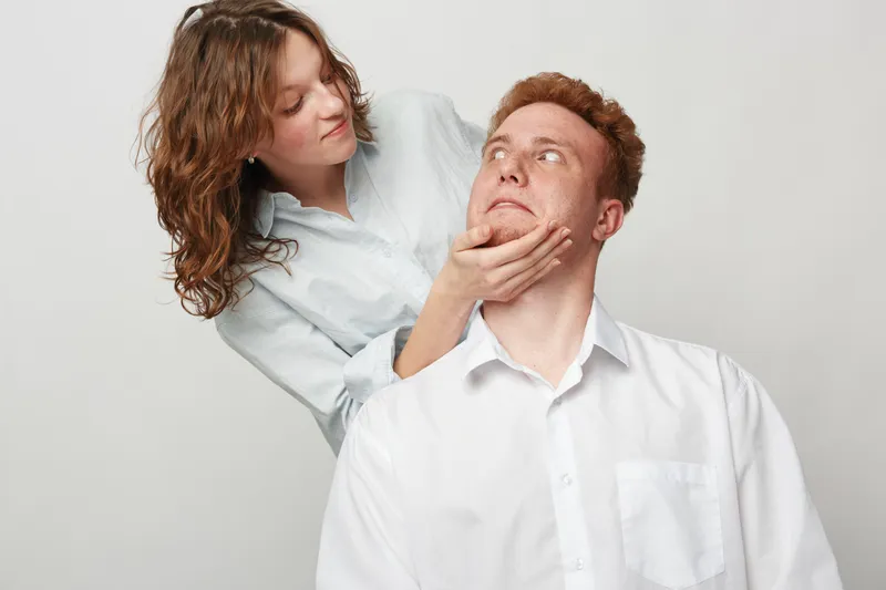 Young woman gently holds man’s face while he looks up with surprised expression. Studio portrait on white background. Concept of relationship, emotion, and human connection.