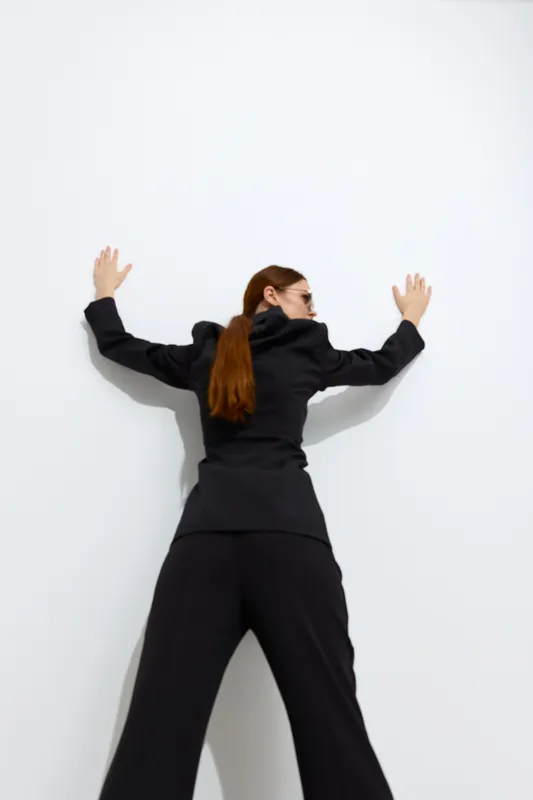 Fashion woman with long red hair wearing a black outfit poses against a white wall, showcasing modern style and minimalistic elegance in a studio setting.