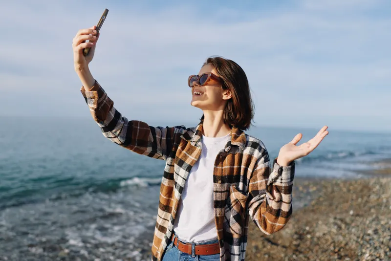 Woman taking a selfie on a rocky beach, wearing sunglasses and a plaid shirt, casual style with jeans, as the ocean and blue sky frame a sunny coastal moment