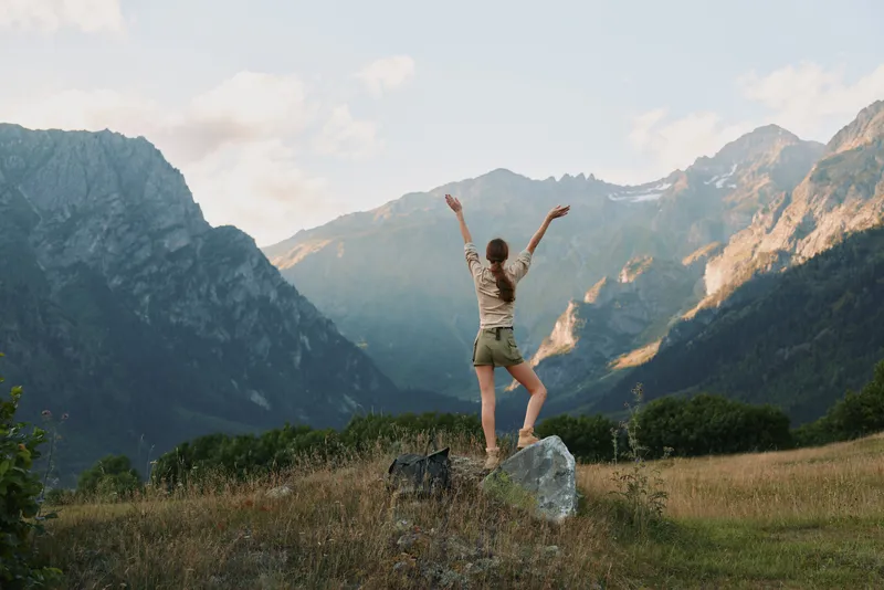 Young woman with backpack stands on rock in mountain valley raises arms enjoys nature and freedom during hiking adventure in scenic landscape outdoors.