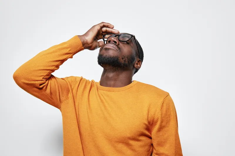 African American man with dark skin wearing glasses and orange sweater touching forehead with eyes closed on white background. Portrait of thoughtful black male in casual clothing.