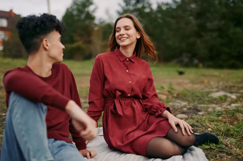 Young couple sitting on a blanket in a grassy park, sharing a relaxed candid moment with natural smiles and subtle gestures that capture an authentic, trustworthy everyday connection.