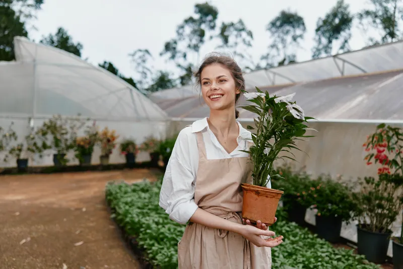 Young woman in an apron holding a potted plant in a greenhouse, showcasing joy and connection to nature in a serene setting