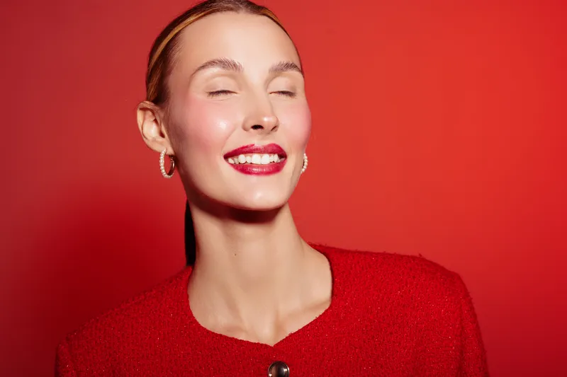 Smiling woman with closed eyes wears red jacket and lipstick, posing on red background. Concept of beauty, fashion, happiness, and confident female model in studio lighting.