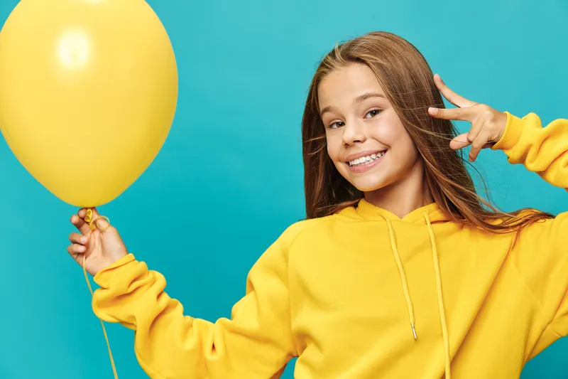 Girl smiling and holding yellow balloon with peace sign wearing yellow hoodie on blue background. Happy child portrait with casual clothes and cheerful expression.