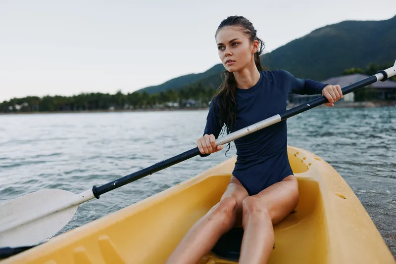 A focused woman sits in a yellow kayak, gripping a paddle as gentle waves touch the shore. She wears a dark blue wetsuit, ready for a calm lake paddle.