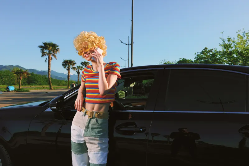 A woman with a blonde curly wig in a striped top leans against a sleek car in a sunny lot, channeling retro lifestyle vibes, bold color, and playful fashion.