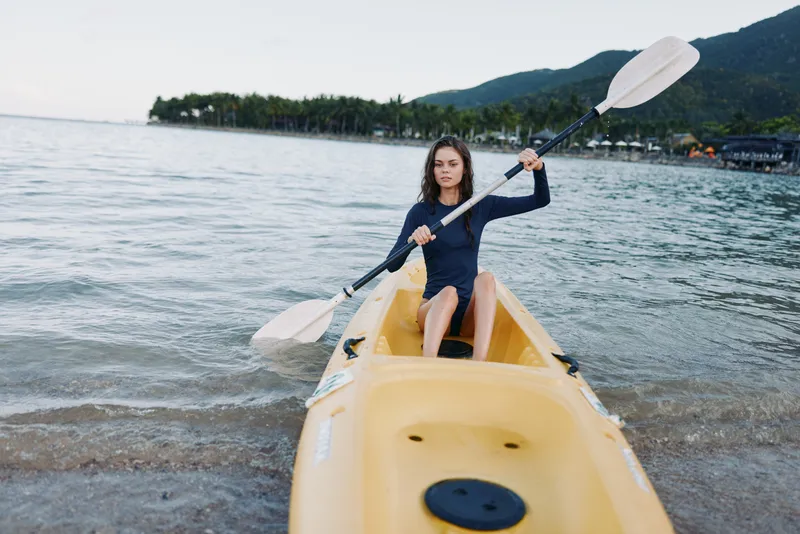 A young paddler steers a bright yellow kayak along a calm shoreline, paddling with focus as gentle waves lap the sand, seabreeze rustling hair, under a blue sky.