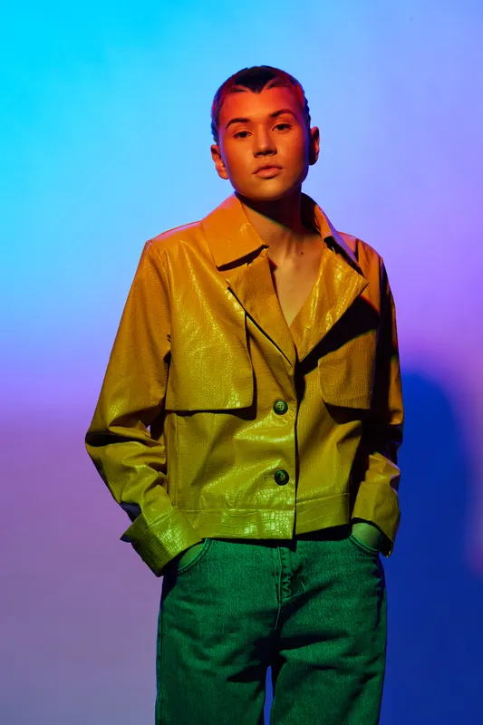 woman with short hair wearing yellow jacket and blue pants, posing confidently against colorful gradient background under vibrant lighting.