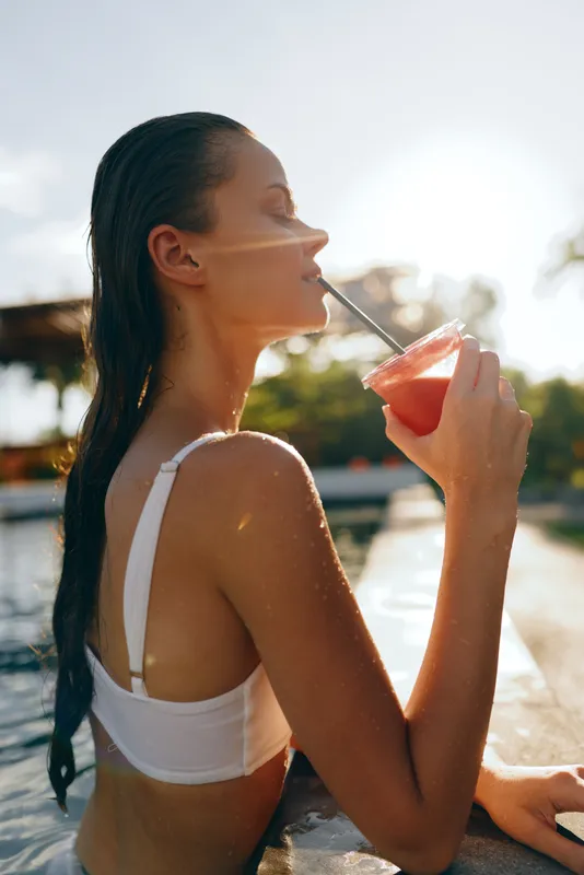 A relaxed woman in a white swimsuit sips a bright beverage by a sunny pool, portraying a casual summer moment of leisure, hydration, and outdoor lifestyle, confident and serene.