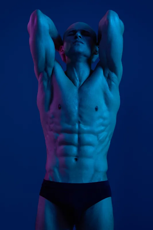 Muscular man posing in dramatic lighting, showcasing toned abs and strong physique, emanating confidence and strength. The vibrant blue and purple hues enhance the visual impact.