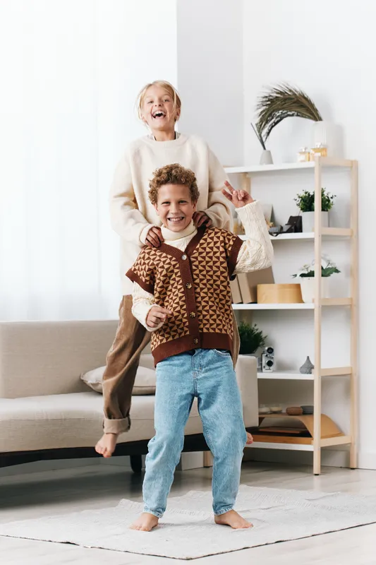 Two joyful children dancing indoors, a smiling boy in denim jeans leads the moment while a girl stands on a sofa behind, creating a bright, playful family scene full of energy.