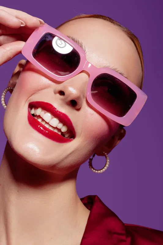 Close-up of smiling woman wearing pink sunglasses and red lipstick against purple background. Fashion portrait with hoop earrings and shiny skin. Concept of style, beauty, and confidence.