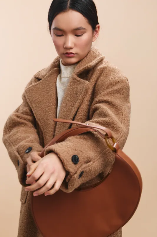 asian woman wearing a cozy camel coat adjusts a brown handbag in a studio portrait, radiating calm confidence, warm textures, and urban fashion appeal in a minimalist setting.