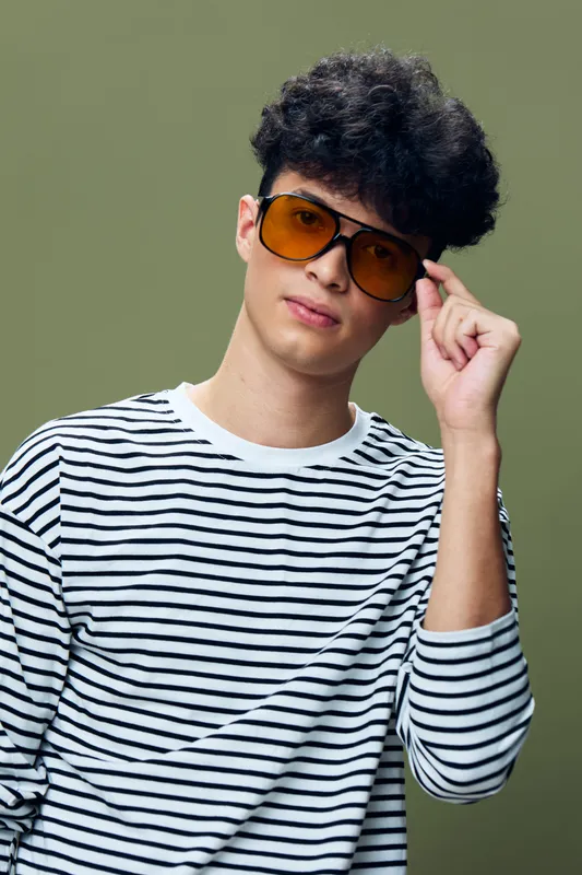young man wearing striped shirt and orange sunglasses posing confidently against green background with curly hair and casual style