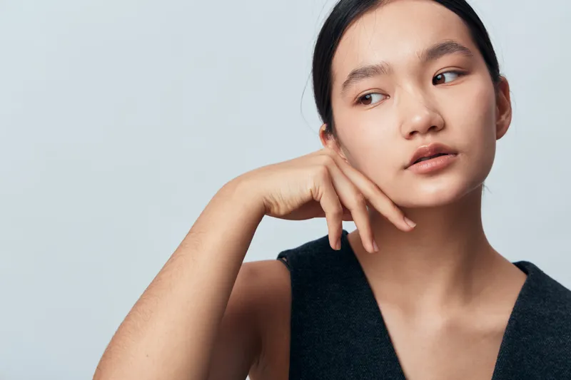 Young woman with clear skin and natural makeup posing thoughtfully with hand near face against light background. Beauty, fashion, portrait, ethnic model with sleek hair and casual black top.