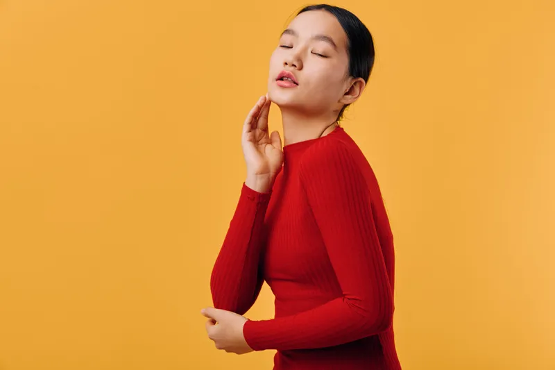 Young woman with eyes closed wearing red sweater standing against orange background. Portrait of peaceful female model posing with hand near face for fashion and beauty concept.