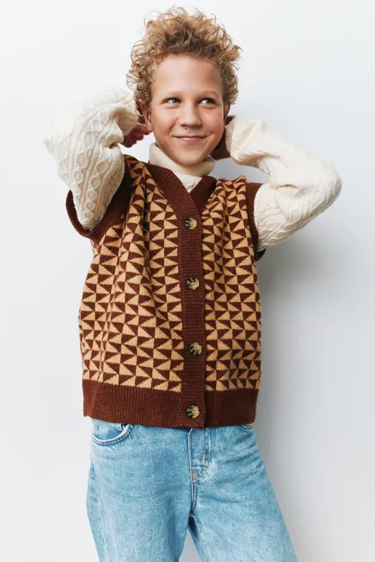 Cheerful young boy poses in a brown geometric knit vest over a creamy sweater, showcasing casual autumn style, confident stance, and a bright, engaging smile.