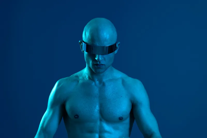 Muscular man with a bald head showcases futuristic fashion, wearing reflective sunglasses and striking a powerful pose in a blue-toned atmosphere.