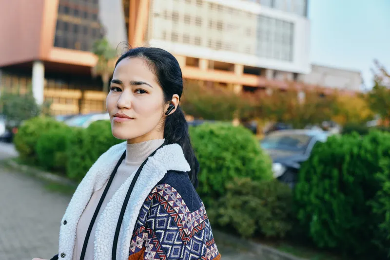 Woman wearing a patterned cardigan and scarf stands outdoors along a street with greenery and buildings in the background, highlighting casual style, urban fashion, and everyday confidence in natural