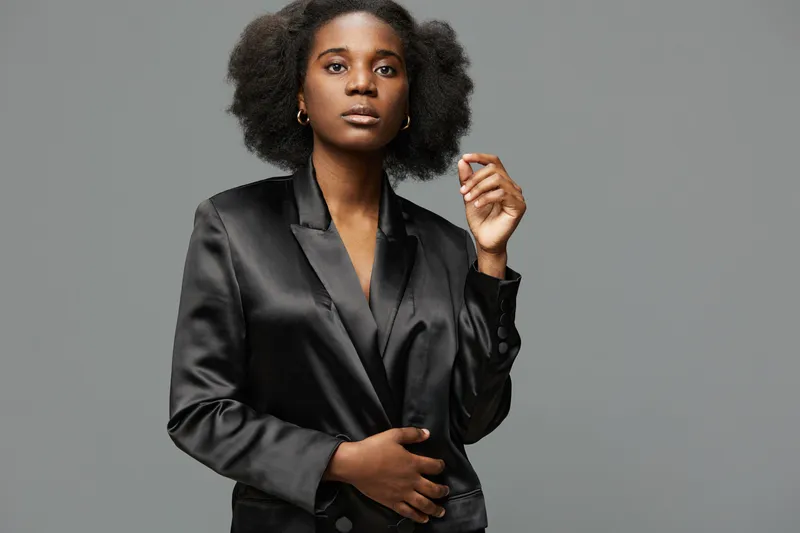 Elegant confident Black woman in a stylish black blazer, showcasing a bold expression against a minimalist gray background, emphasizing modern professional fashion