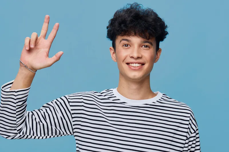 Smiling man in striped shirt making peace sign against blue background, young and casual with curly hair, cheerful and friendly expression.