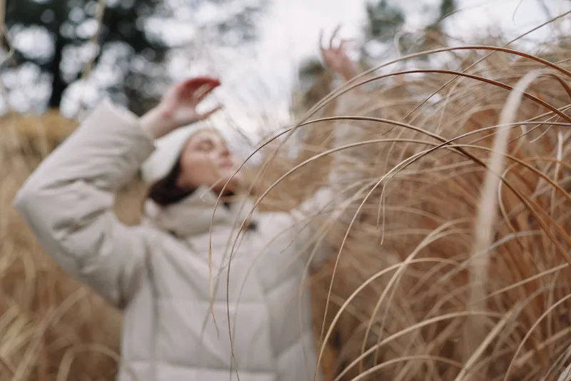 Outdoor scene with a lone figure in a puffy coat standing in a tall grass field, wind sweeping through the blades as the person shields their face and explores quiet, natural textures and winter