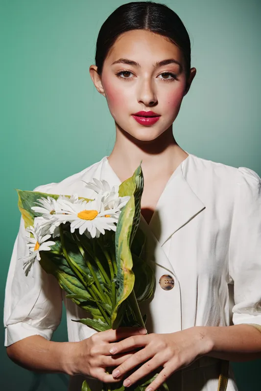 Elegant young woman with a stylish white dress holds a bouquet of white daisies against a pale green background. Beauty and fashion concept with clear, detailed lighting.