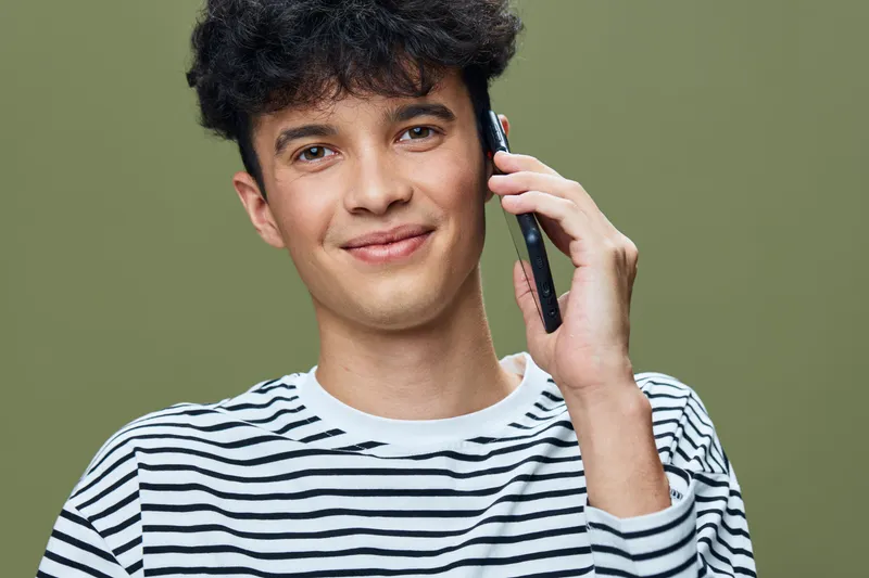 young man talking on smartphone wearing striped shirt with curly hair in front of green background, casual communication and modern lifestyle portrait