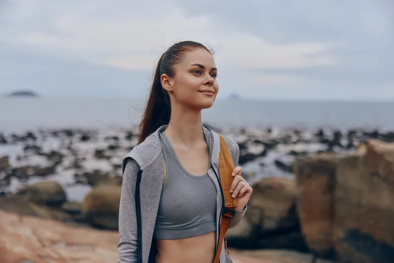 Young woman in athletic attire smiles confidently by the seaside, wearing a grey sports bra and hoodie, perfect for fitness and healthy lifestyle concepts