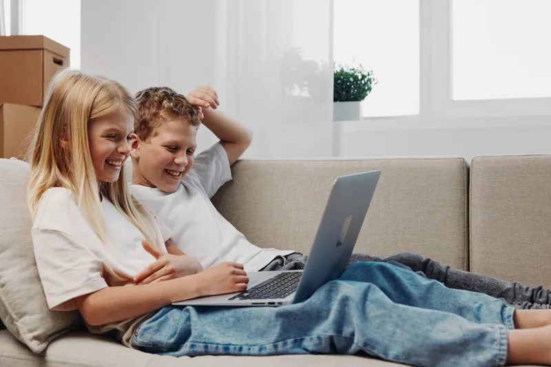 Two happy children sitting on a couch using a laptop, smiling and enjoying digital entertainment at home in a bright living room.