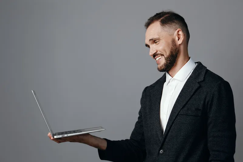 A professional man in a suit smiles, holding a laptop on his palm against a neutral colored background, projecting confidence, warmth, and an approachable business mood.