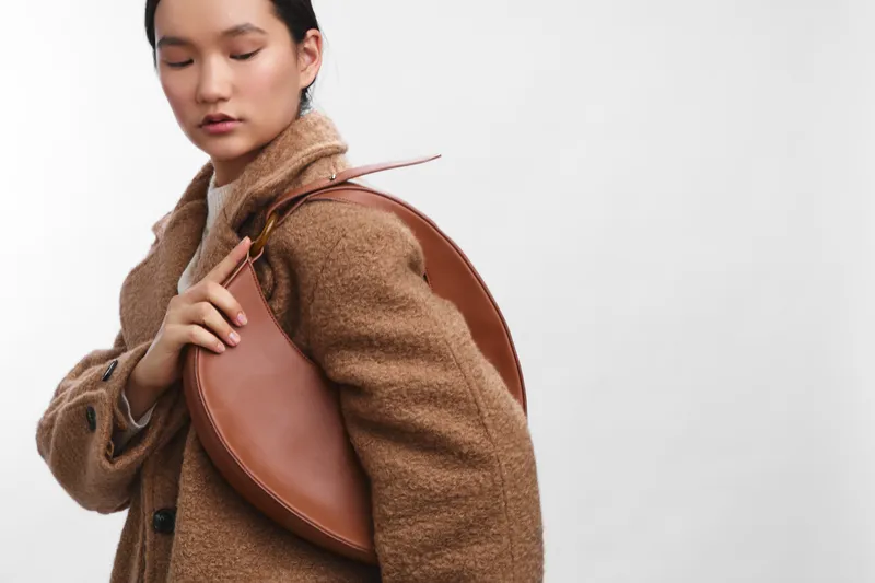 Asian woman wearing a tan coat stands with a circular bag in a clean studio, creating a calm, confident fashion moment that highlights contemporary style and versatile outerwear.