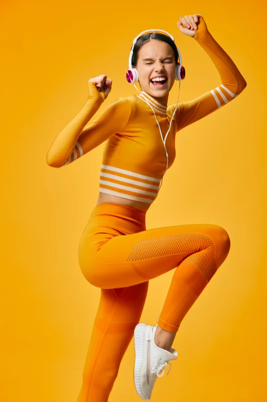 Fitness woman dancing in a bright orange athletic outfit, headphones on, mid jump with energetic rhythm in a bold studio. Dynamic movement, sport performance, and vibrant lifestyle vibe for fitness