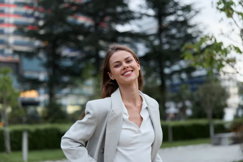 Young woman smiling confidently outdoors in casual business attire with modern urban background, natural light, and relaxed pose KEYWORDS: woman smiling, business casual, urban background, confident y