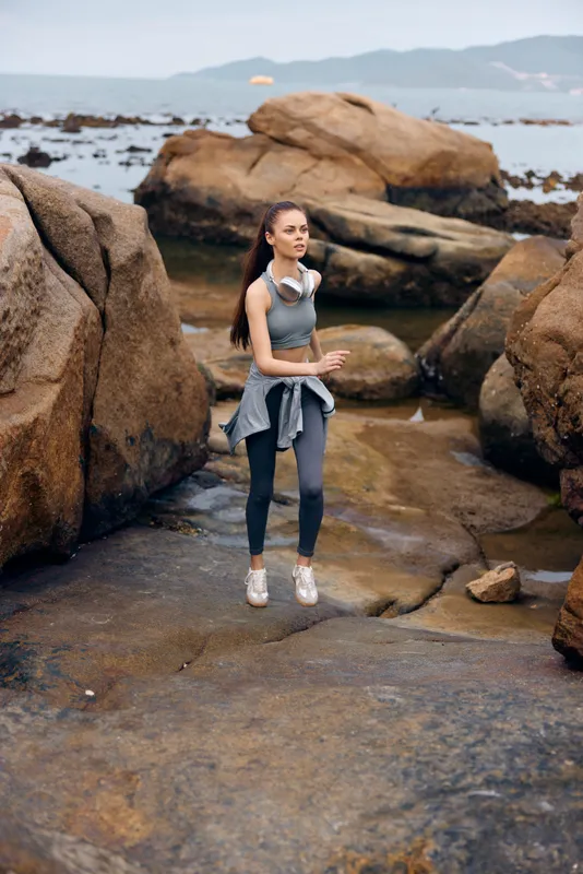 Young woman jogging on rocky shoreline wearing sportswear, headphones, with a determined expression, embodying fitness and outdoor lifestyle concepts