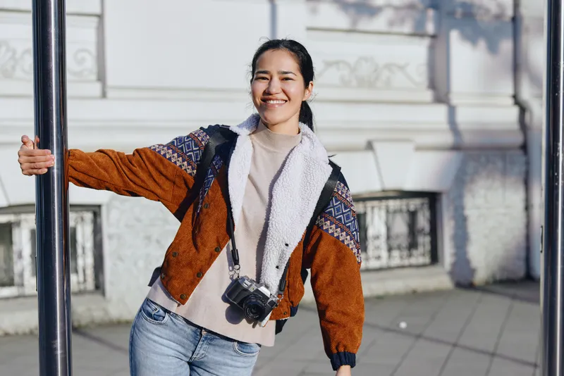 Woman in a warm brown jacket with a scarf and camera around her neck, smiling outdoors on a city street, casual fashion and photography moment in an urban setting