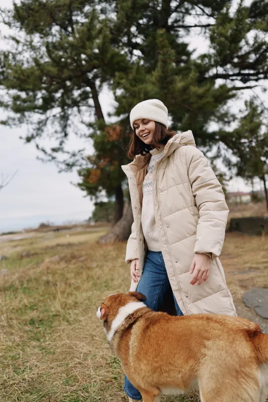 Woman in a beige winter coat and beanie smiles as she interacts with a dog outdoors in a parklike setting with trees and autumn tones.