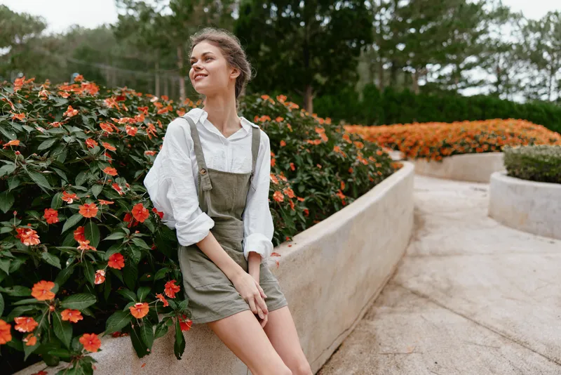 Young woman in green overalls with white shirt smiling near vibrant orange flowers in a sunny garden showcasing a joyful spring atmosphere