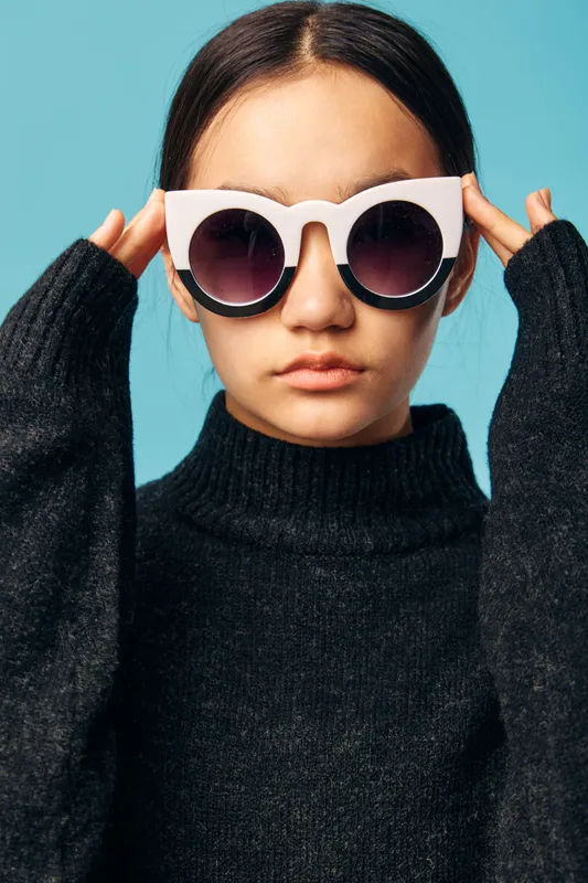 Young woman wearing oversized white sunglasses and black turtleneck sweater poses against blue background with serious expression and hands adjusting frames near face.