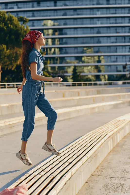 lifestyle woman in modern boho-western denim street style jumping outdoors, casual outfit with red bandana and sneakers, film color tones capturing urban energy.