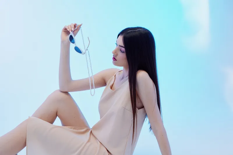 Young woman with long dark hair in a beige dress holds sunglasses and a pearl necklace while sitting. Studio shot with soft blue background. Concept of woman beauty fashion.