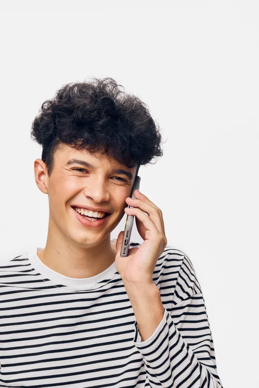 smiling young man wearing striped shirt talking on smartphone with curly hair against white background, cheerful expression, casual communication moment