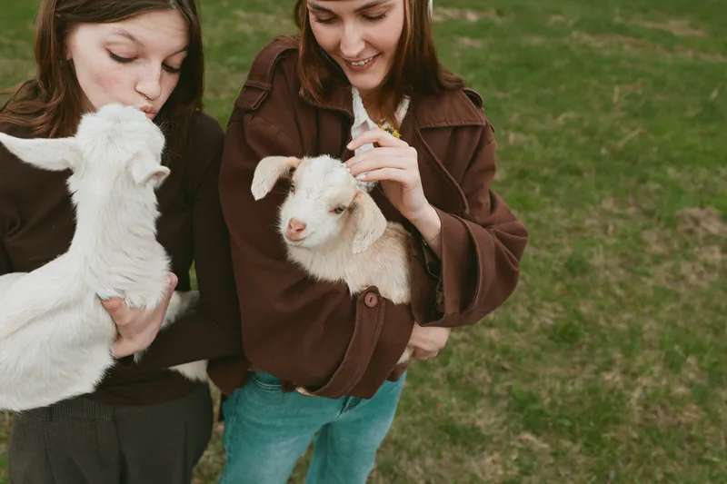 Two young women on a grassy field interact with friendly goats, one cradling a kid while the other smiles, presenting a warm casual lifestyle moment with gentle affection.